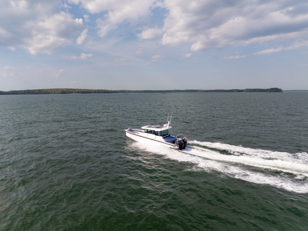 Aerial view of an Axopar 29 XC Cross Cabin with a white hull cruising across open water, leaving a long white wake under a partly cloudy sky.