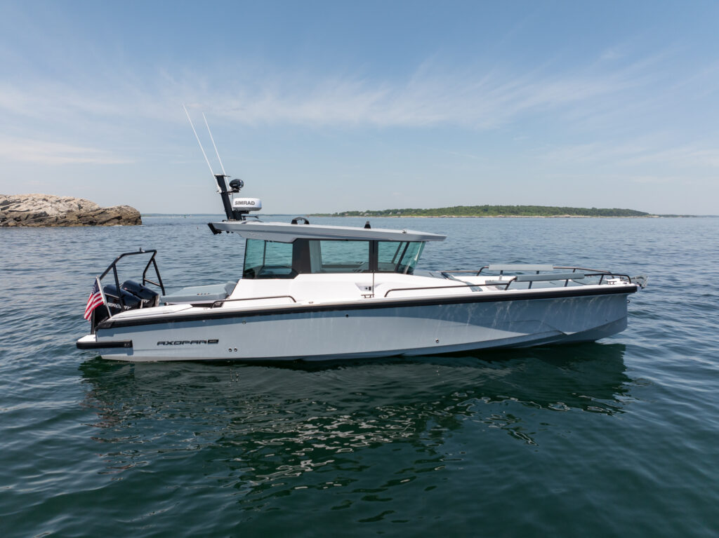 Side profile of a platinum grey BRABUS Axopar 29 XC Cross Cabin floating on calm water, with a rocky outcropping and a low green island in the distance.