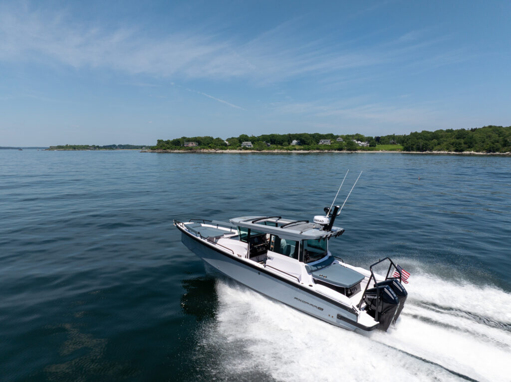 Aerial view of a BRABUS Axopar 29 XC Cross Cabin with a platinum grey hull cruising at speed on calm water, leaving a wide white wake with a green shoreline in the background.