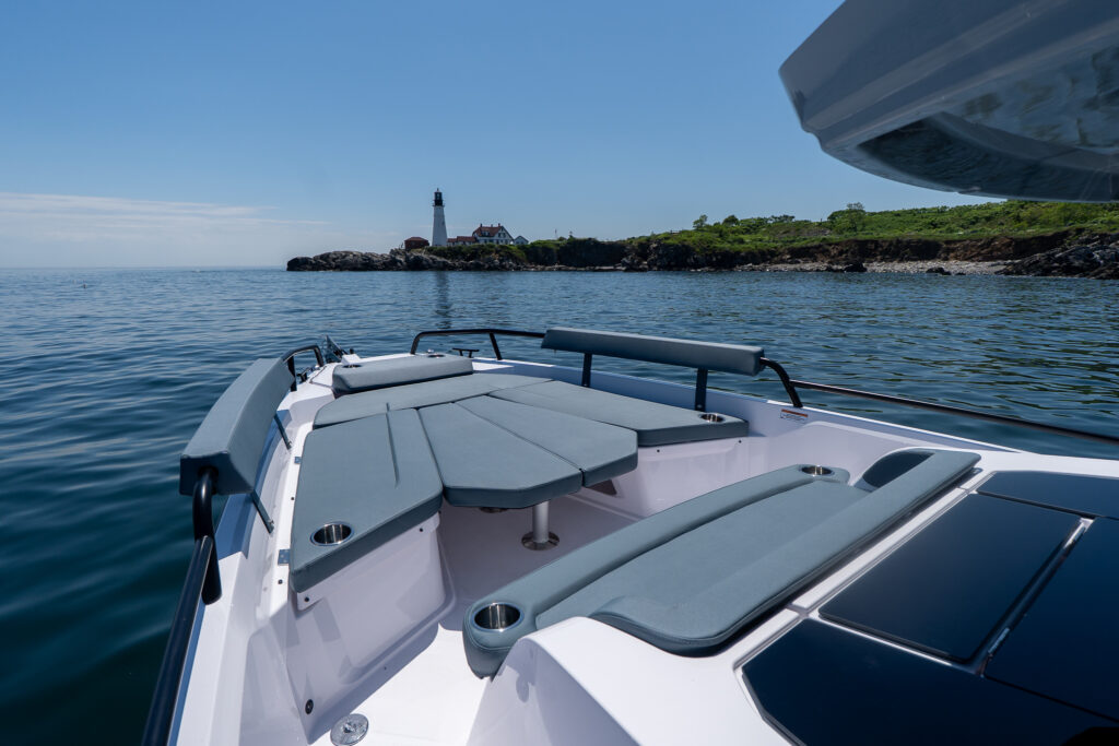 Axopar 29 XC Cross Cabin BRABUS Edition bow seating and sun lounge in Petrol upholstery with cupholders, with Portland Head Light on the rocky shoreline ahead.