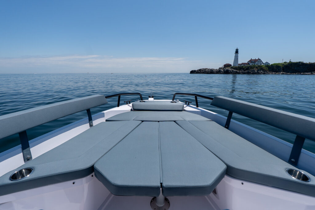 Bow sun pad with Petrol upholstery and side backrests on an Axopar 29 XC Cross Cabin BRABUS Edition, with Portland Head Light in the distance across calm water.
