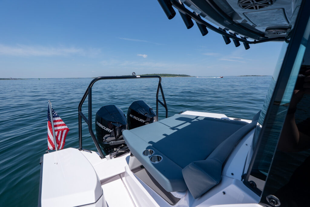 Aft deck view from the Axopar 29 XC Cross Cabin BRABUS edition, showing twin black Mercury outboards, a black BRABUS ski-tow bar, an American flag, and a Petrol-upholstered aft sun pad with cupholders on calm blue water.