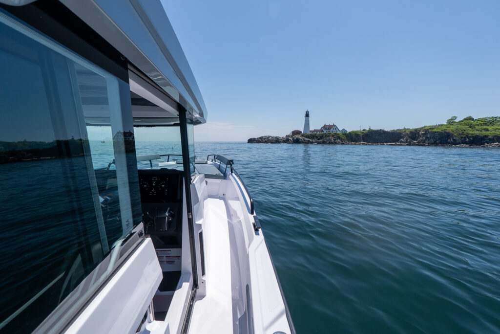 Starboard-side walkway of an Axopar 29 XC Cross Cabin running past the pilothouse windows, with calm water and Portland Head Light ahead on a rocky point.