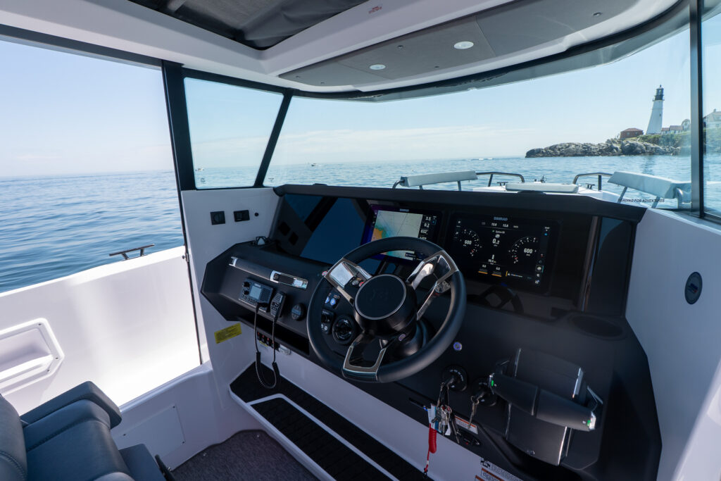 Angled view of the Axopar 29 XC Cross Cabin BRABUS edition helm with BRABUS steering wheel, twin Simrad displays, and throttle controls, with Portland Head Light visible through the windshield on the right.