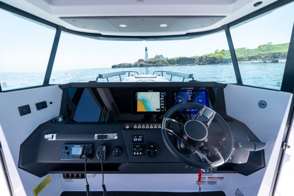 Wide view of the Axopar 29 XC Cross Cabin BRABUS edition helm with twin Simrad displays and BRABUS steering wheel, looking out through the windshield toward Portland Head Light on the rocky shoreline.