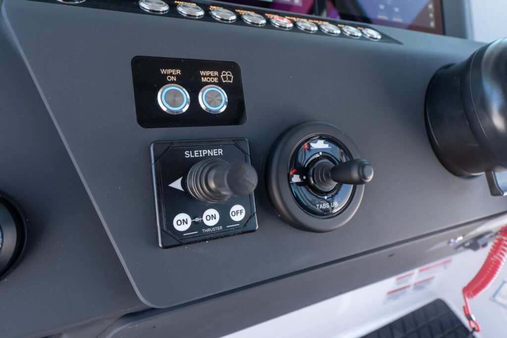 Close-up of the Axopar 29 XC Cross Cabin helm dashboard showing illuminated wiper buttons, a Sleipner thruster joystick, and a trim-tab control lever.