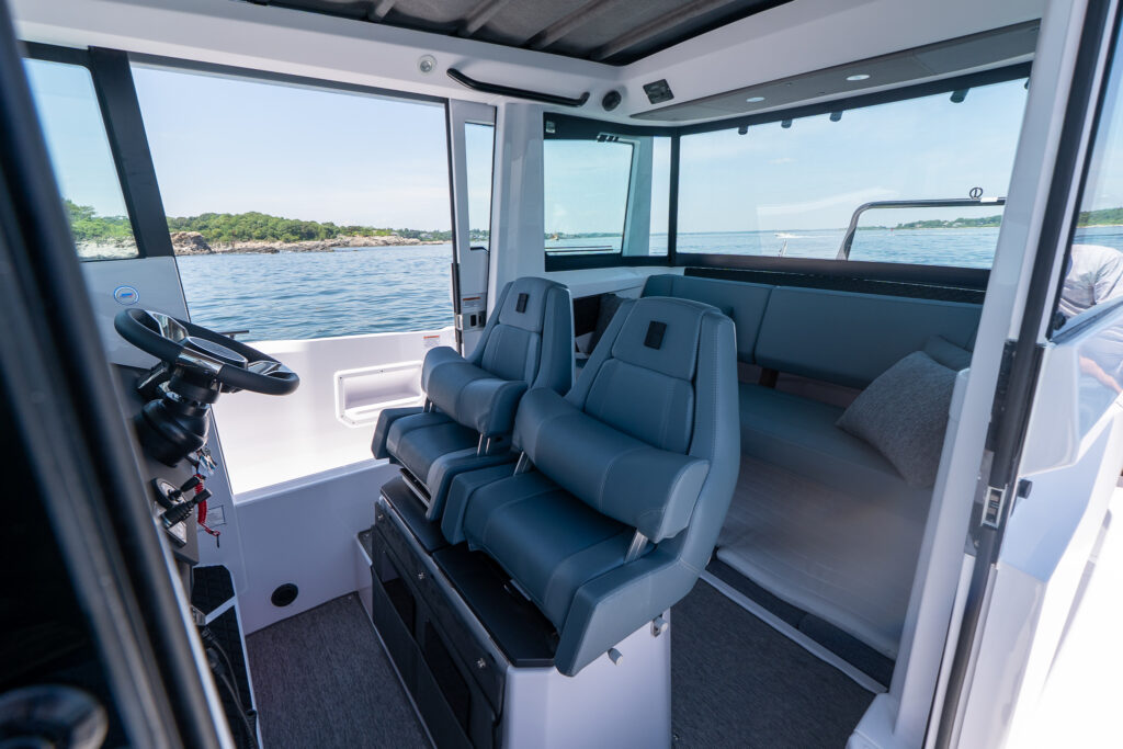Pilot house view showing two Petrol-upholstered helm seats with the flip-up bolsters raised, positioned in front of the helm, with an aft bench and large windows looking out over calm water.