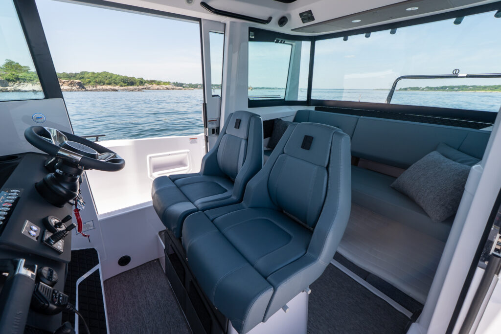 Pilot house interior with two Petrol-upholstered helm seats facing a steering wheel and controls, plus an aft bench with pillows beside large windows overlooking calm water.