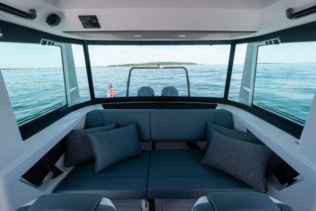 Enclosed aft cabin seating area with dark blue cushions and throw pillows, looking out through wraparound windows toward the ski tow bar and twin Mercury outboards on calm water.