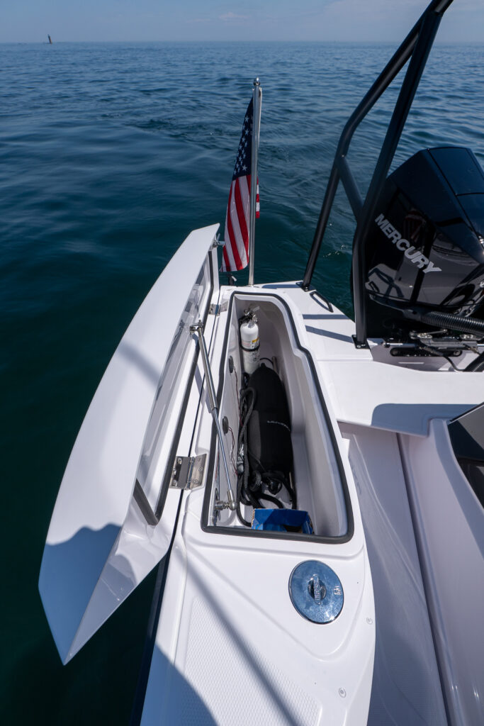 Open fender locker at the stern with a fire extinguisher and stowed gear inside, next to the black BRABUS ski tow bar and a Mercury outboard, with an American flag flying and Halfway Rock visible far on the horizon.