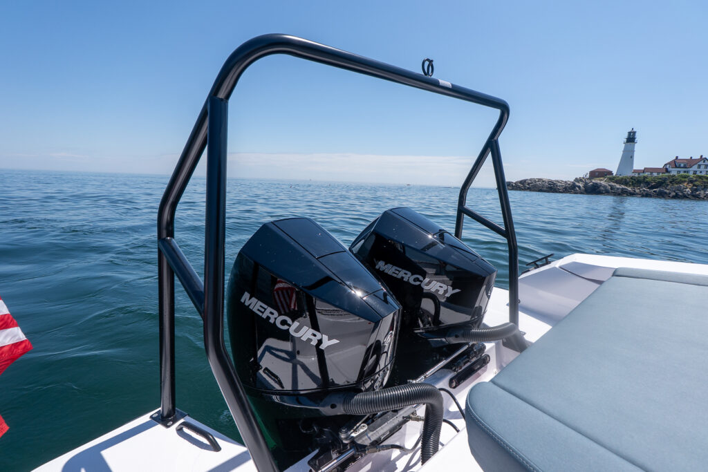 Close-up of a black BRABUS ski tow bar framing twin black Mercury outboards at the stern, with a Petrol upholstery sunpad in the foreground and Portland Head Light on the rocky shoreline in the distance.