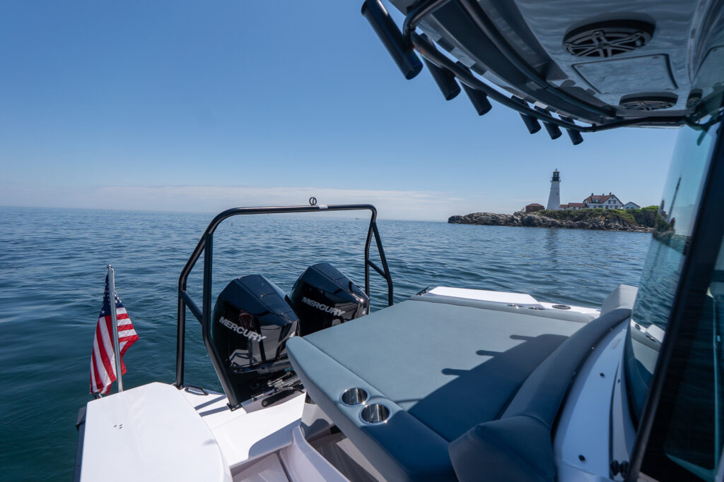 Aft deck view of a BRABUS edition Axopar 29 XC Cross Cabin with a black ski tow bar, Petrol upholstery sunpad, twin black Mercury outboards, and Portland Head Light off the starboard side.