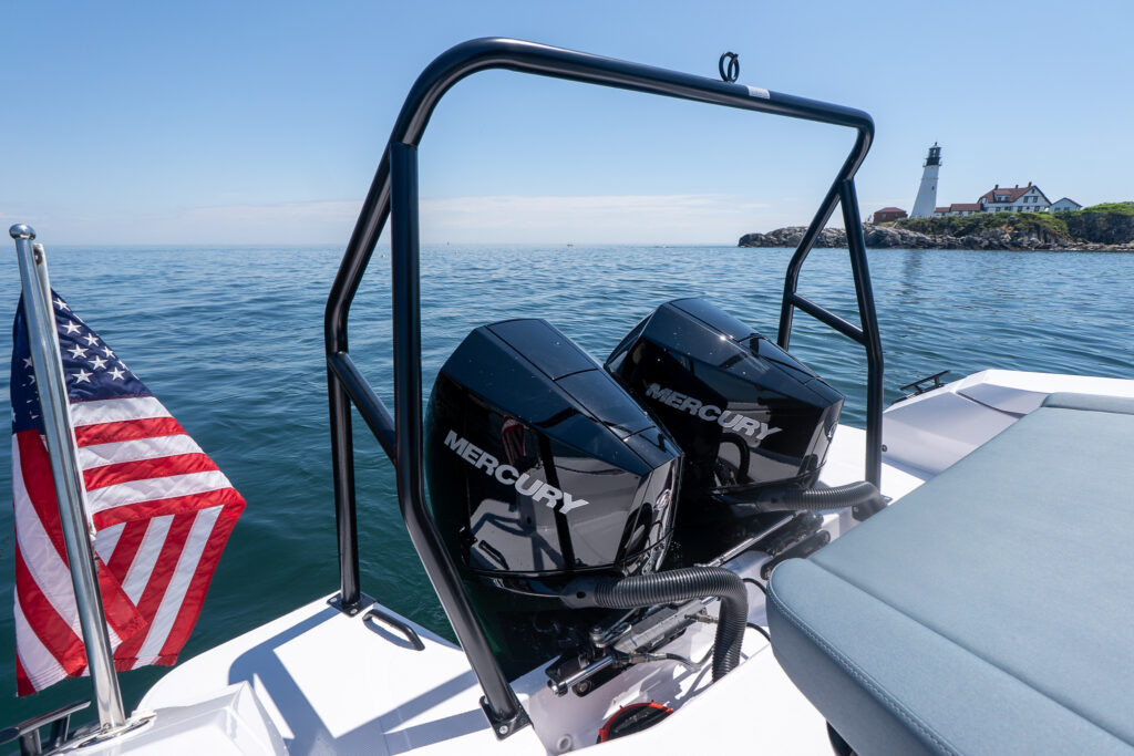 Twin black Mercury outboard engines beneath a black BRABUS edition ski tow bar on an Axopar, with an American flag at the stern and Portland Head Light in the distance.