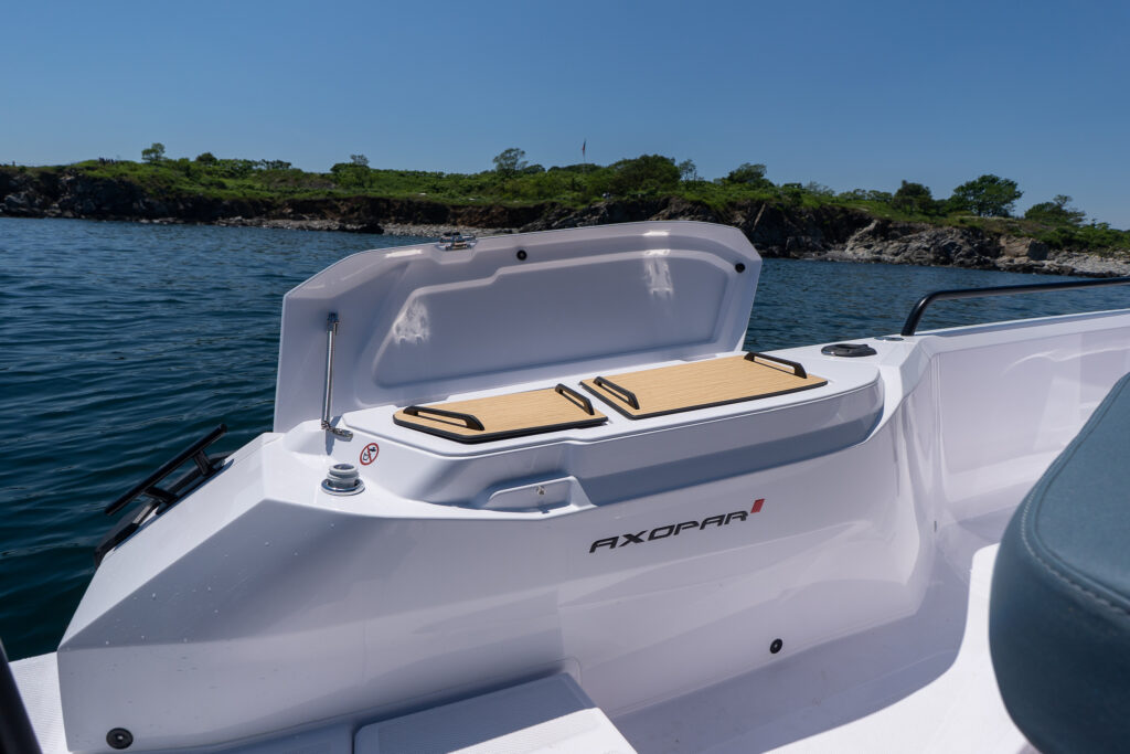 Open fender locker wet bar on an Axopar 29 XC Cross Cabin, showing covered bar inserts and the Axopar logo, with calm water and a rocky green shoreline behind.