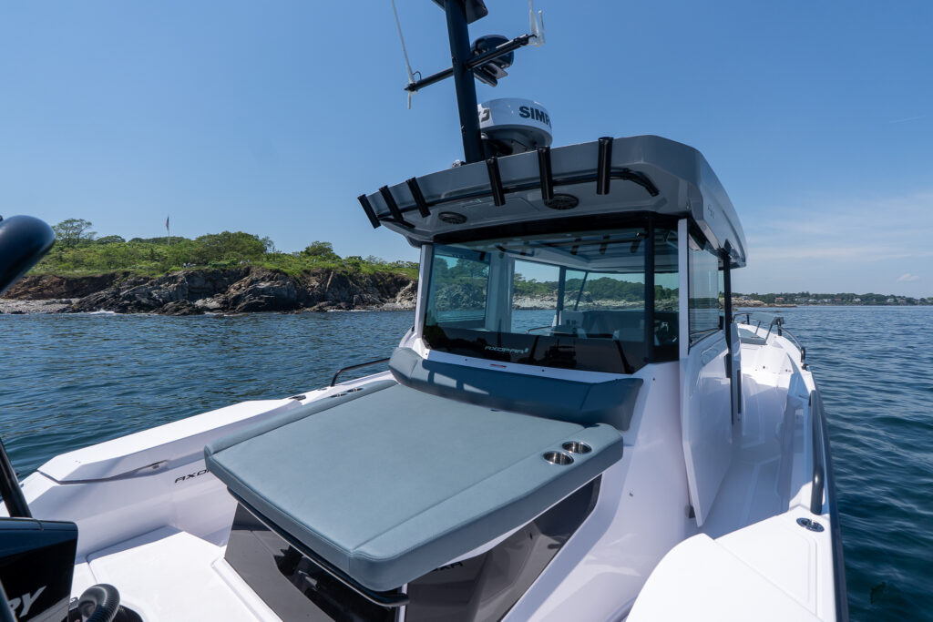 Aft cabin sunpad with Petrol upholstery on a BRABUS Edition Axopar 29 XC Cross Cabin, showing rod holders, radar, and pilothouse while floating near a rocky Maine shoreline.