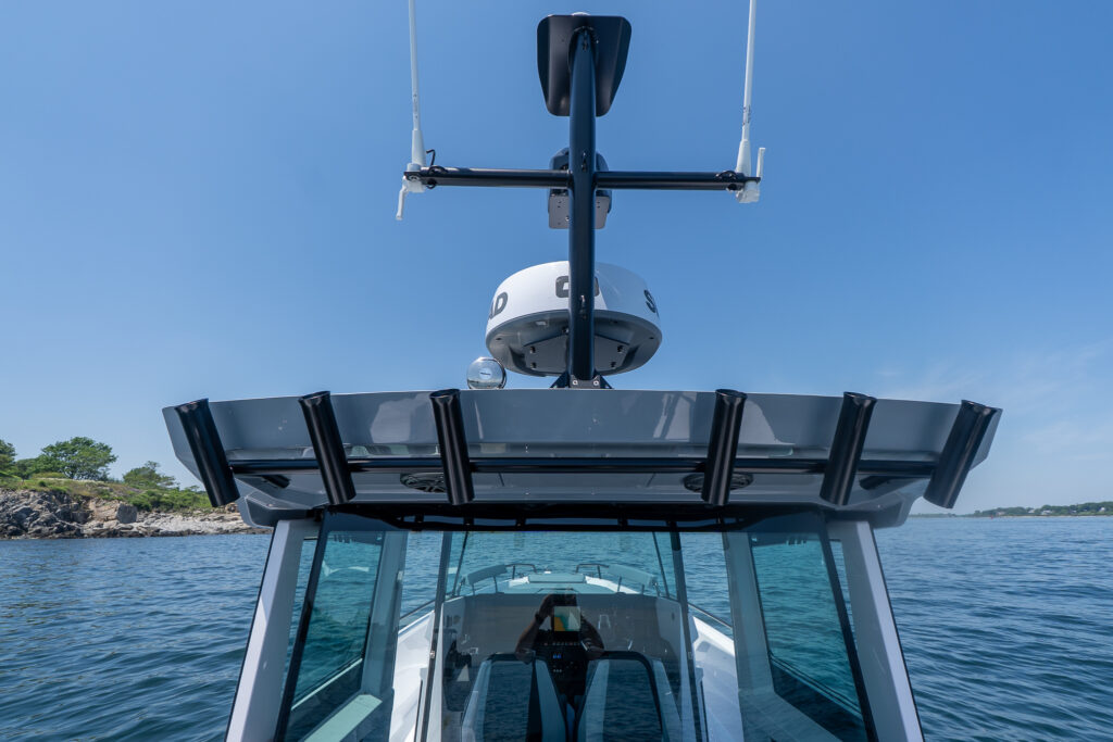 Close-up view of the Axopar 29 XC Cross Cabin’s roof showing rod holders, radar, antennas, and spotlight, with the pilothouse and water in the background.