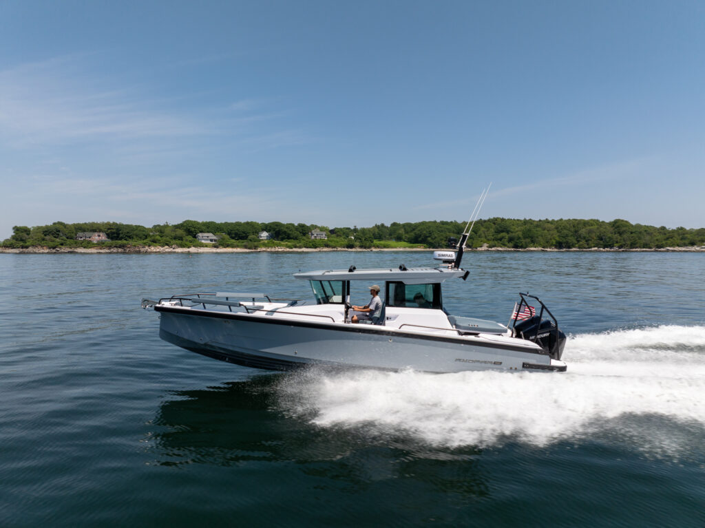 Side profile of an Axopar 29 XC Cross Cabin cruising on plane with a visible wake, helm occupant, and a wooded shoreline in the background.
