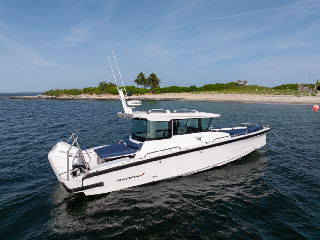 Axopar 29 XC Cross Cabin cruising near a sandy shoreline in Casco Bay, Maine, showing Sphere-upholstered aft seating and a single Mercury V10 Verado.