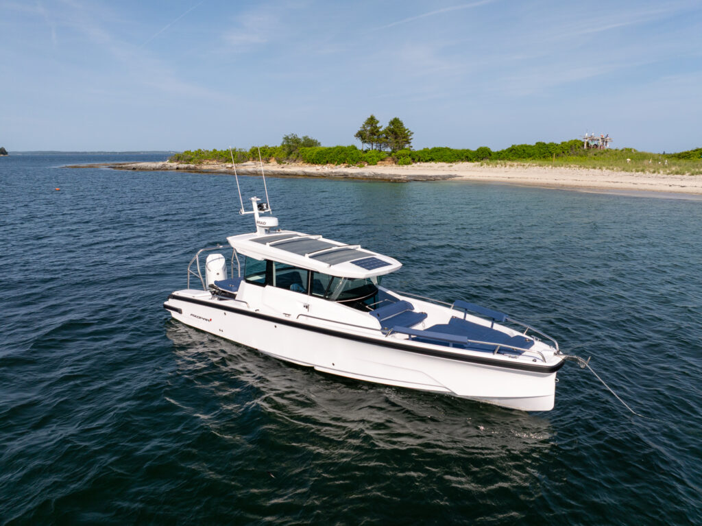 Axopar 29 XC Cross Cabin at anchor near a sandy shoreline in Casco Bay, Maine, showing Sphere-upholstered bow seating and solar panels on the roof.