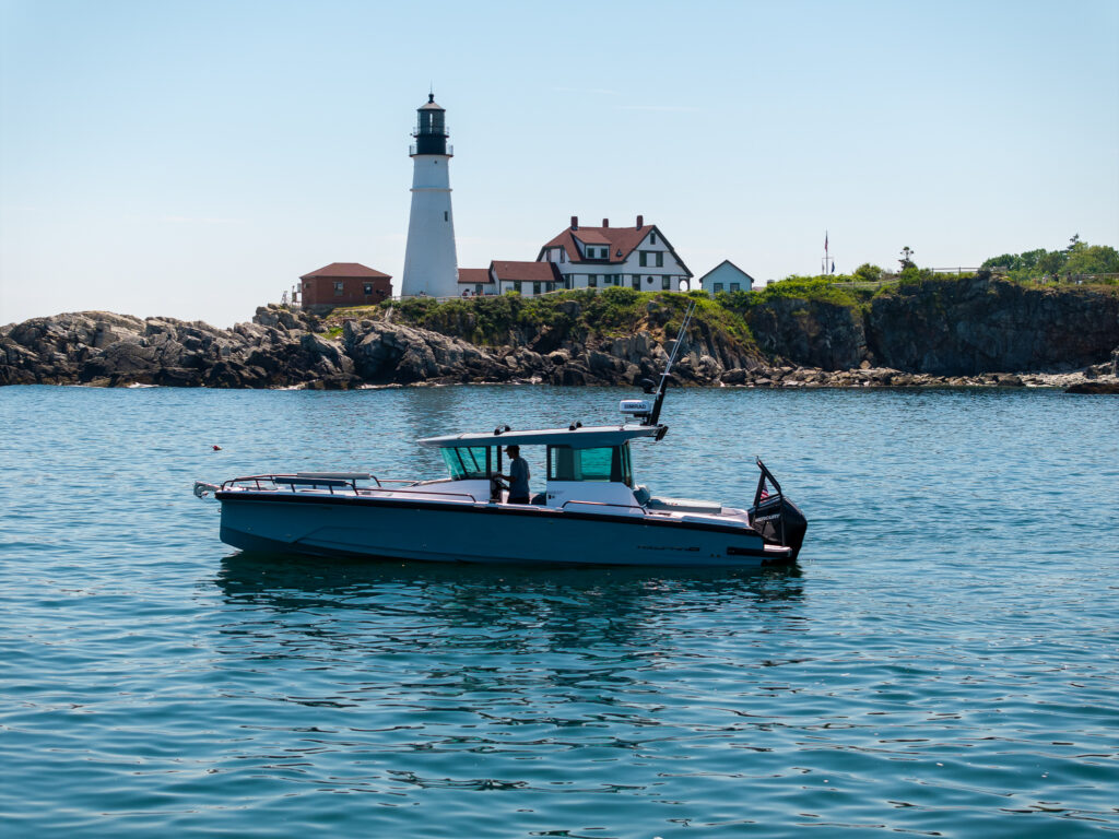 Axopar 29 XC Cross Cabin floating on calm water with Portland Head Light and the keeper’s buildings on the rocky shoreline behind.