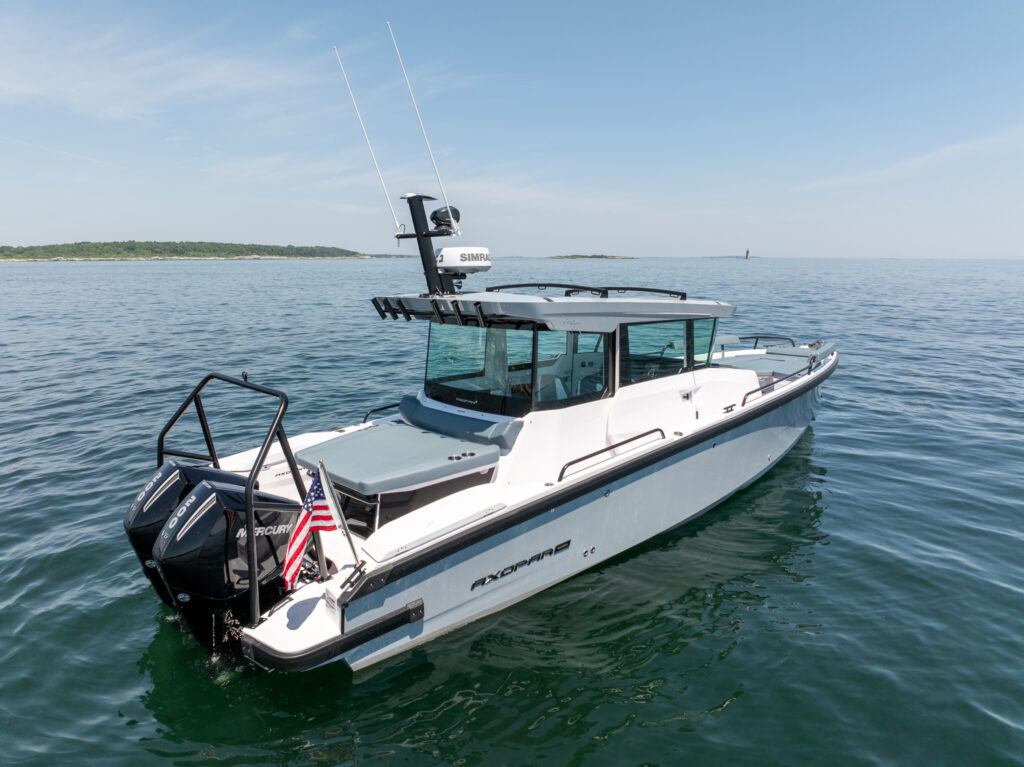 Rear three-quarter view of a BRABUS edition Axopar 29 XC Cross Cabin with a platinum grey hull, twin Mercury outboards, and an American flag on calm coastal water.