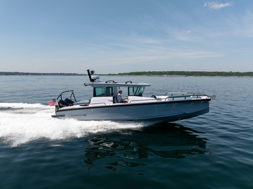 Side profile of a BRABUS Axopar 29 XC Cross Cabin with a platinum grey hull cruising on calm water, leaving a white wake, with a driver at the helm and an American flag at the stern.