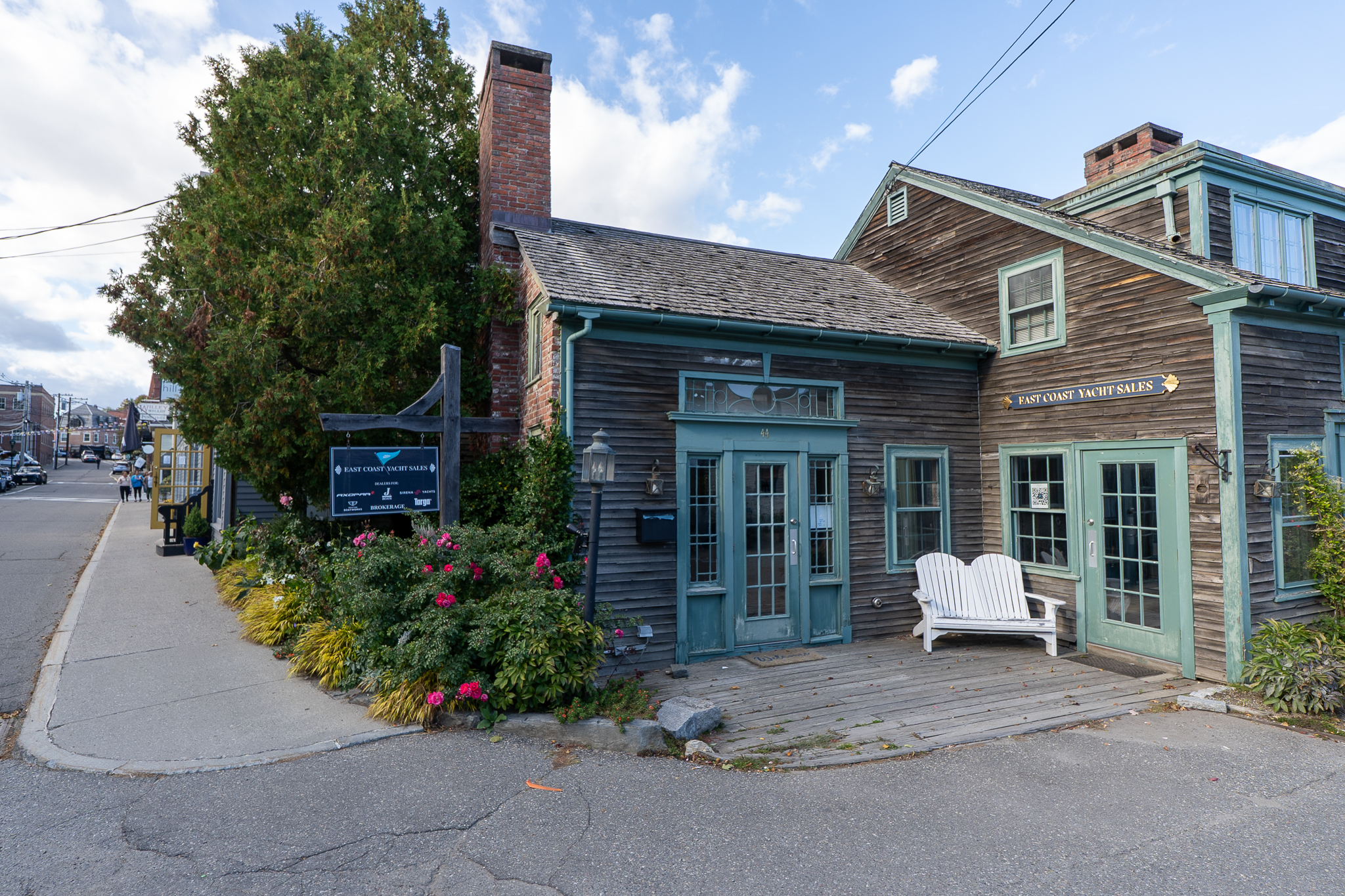East Coast Yacht Sales Camden office on a sunny day with brown wood siding, seafoam green trim, and a white bench out front on a busy Camden, Maine street.