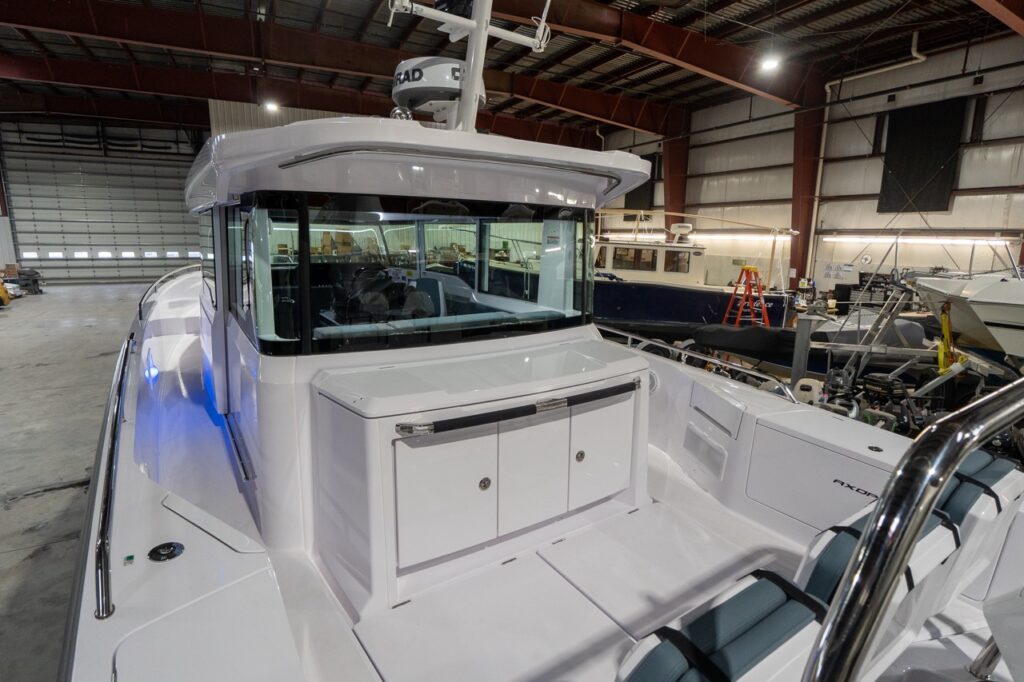Aft deck of an Axopar 37 XC Cross Cabin showing the aft wet bar module behind the pilothouse, with integrated counter space and double cabinet doors, plus staple-style seating with petrol-colored upholstery in the foreground.