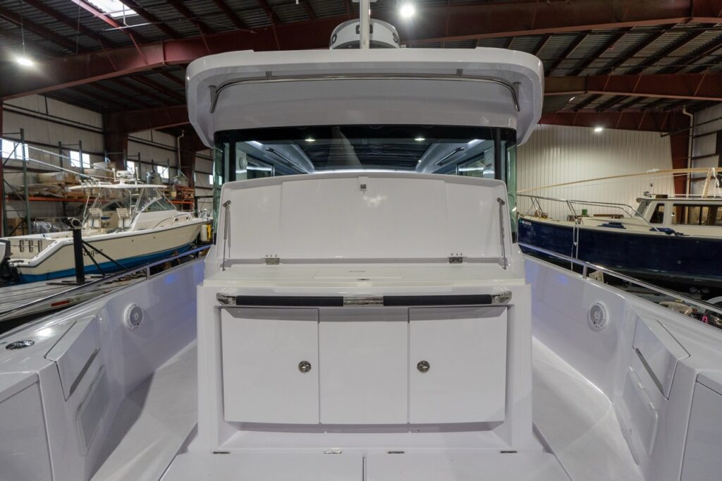 Centered view of the Axopar 37 XC aft wet bar on the aft deck with the upper lid raised, revealing a broad prep surface with an inset hatch, plus double-door storage below and side decks running along both sides.