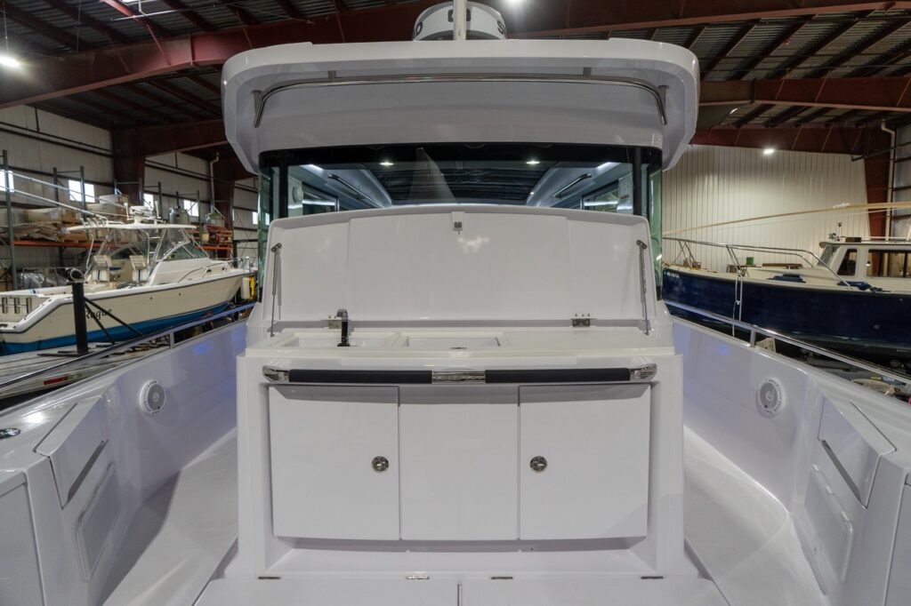 Straight-on view of the Axopar 37 XC aft wet bar on the aft deck with the lid raised, revealing a sink and faucet beside an inset worktop, plus double-door storage below and side decks running along both port and starboard.