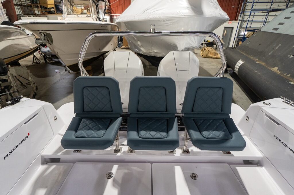 Rear-facing view on the aft deck of an Axopar 37 XC showing three individual staple seats in petrol-colored, diamond-stitched upholstery mounted across the transom, with twin outboard engines and a stainless grab rail behind.