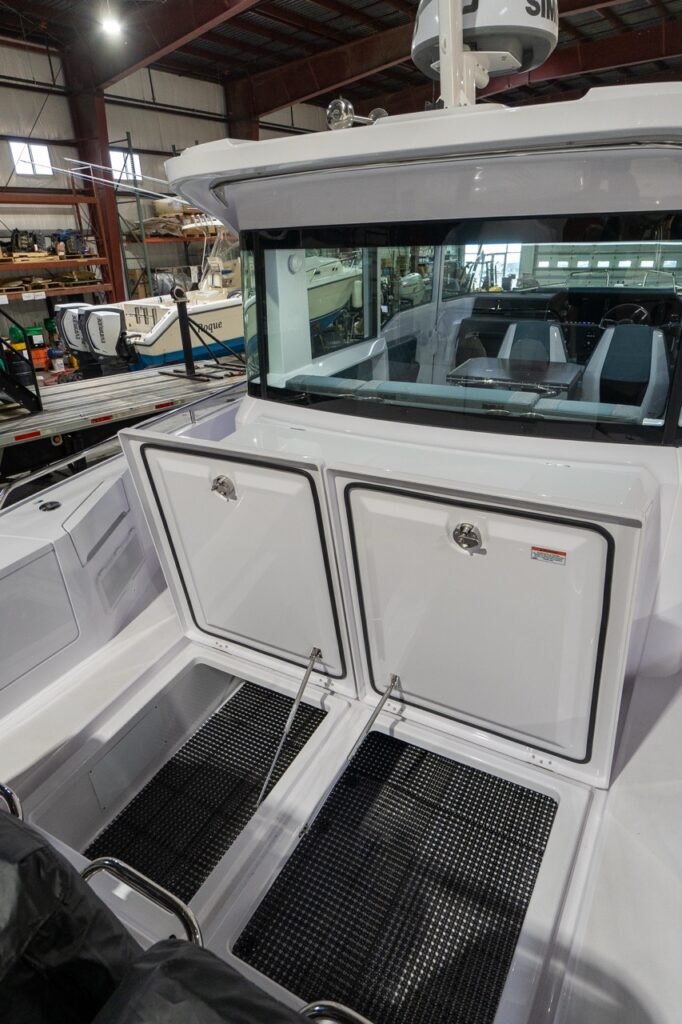 Aft deck view on an Axopar 37 showing two large, gasketed storage locker lids opened aft of the wet bar, revealing deep compartments with non-slip black matting inside, set below the pilot house rear window.