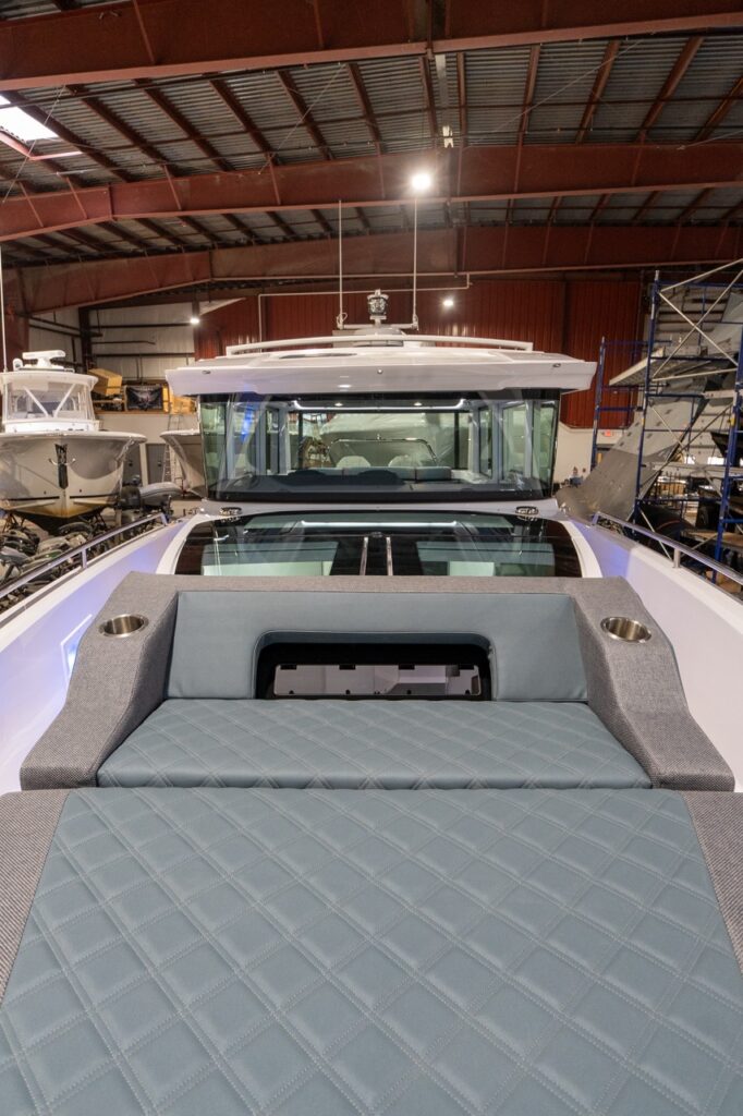 View from the forward deck looking aft on an Axopar 37 XC, showing the quilted petrol sunbed cushions in the foreground and the forward-cabin glass roof formed by the closed gullwing doors, with the pilothouse windshield and roofline beyond.