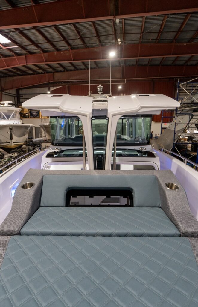 View from the forward deck looking aft on an Axopar 37 XC, with the petrol-upholstered sunbed in the foreground and the twin gullwing doors raised, opening over the forward-cabin skylight area and framing the pilothouse windows behind.