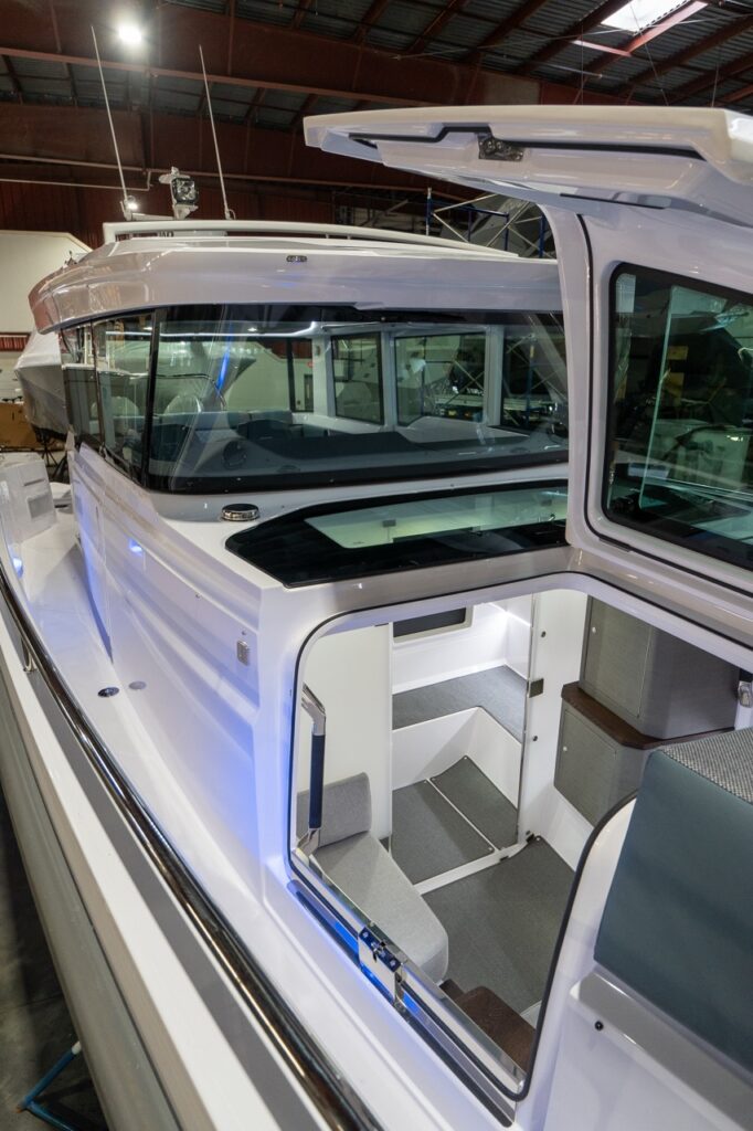 Angled view along the port-side walkway of an Axopar 37 XC with the gullwing door raised, looking down through the opening into the forward-cabin entry steps and the doorway leading to the separate head compartment.
