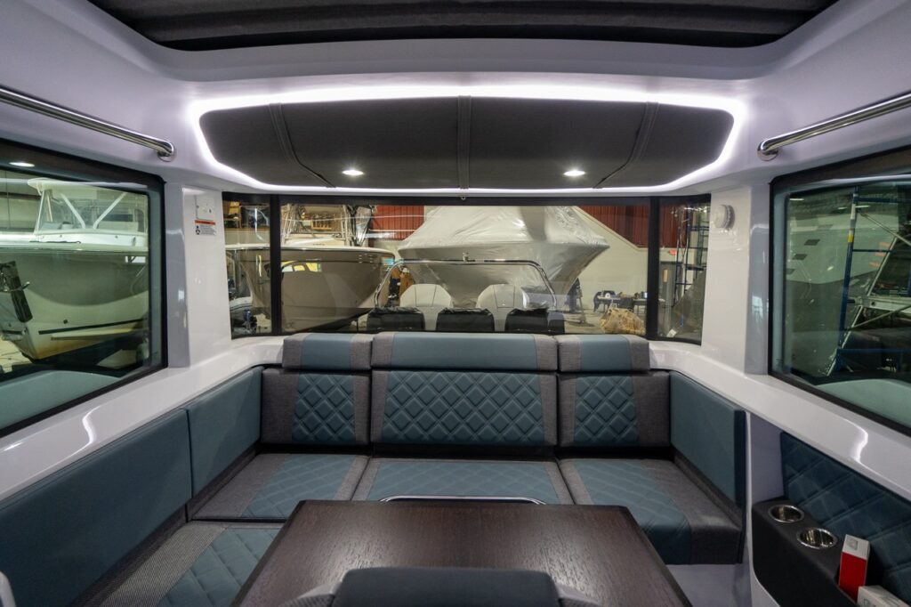 Aft-facing view inside the Axopar 37 XC pilothouse showing the U-shaped aft lounge upholstered in Petrol gray-blue with quilted seat backs, a wood table in the foreground, and a wraparound LED-lit headliner.