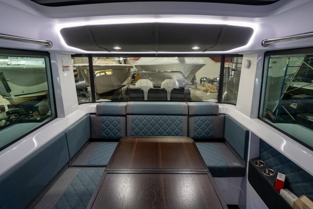 Inside the Axopar 37 XC pilothouse, a U-shaped aft lounge in Petrol gray-blue upholstery wraps around an expandable dark-wood table, with quilted backrests, stainless grab rails, and a halo LED-lit headliner overhead.