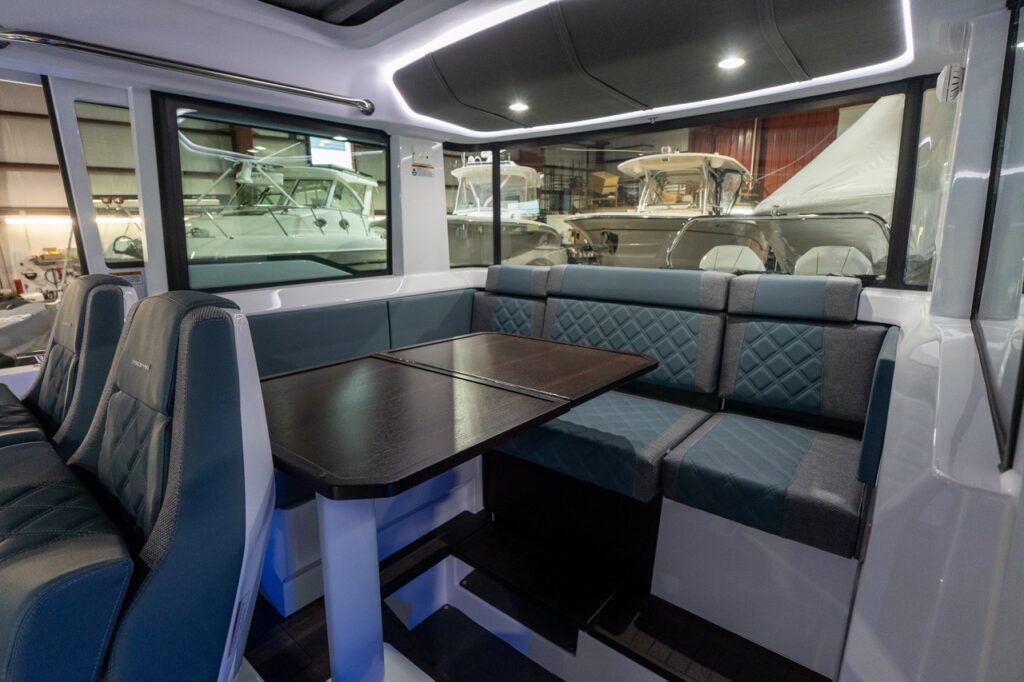 Angled view inside the Axopar 37 XC pilothouse showing the U-shaped aft dinette in Petrol gray-blue upholstery with diamond-stitched backrests, a dark-wood table, and soft LED perimeter lighting in the headliner.