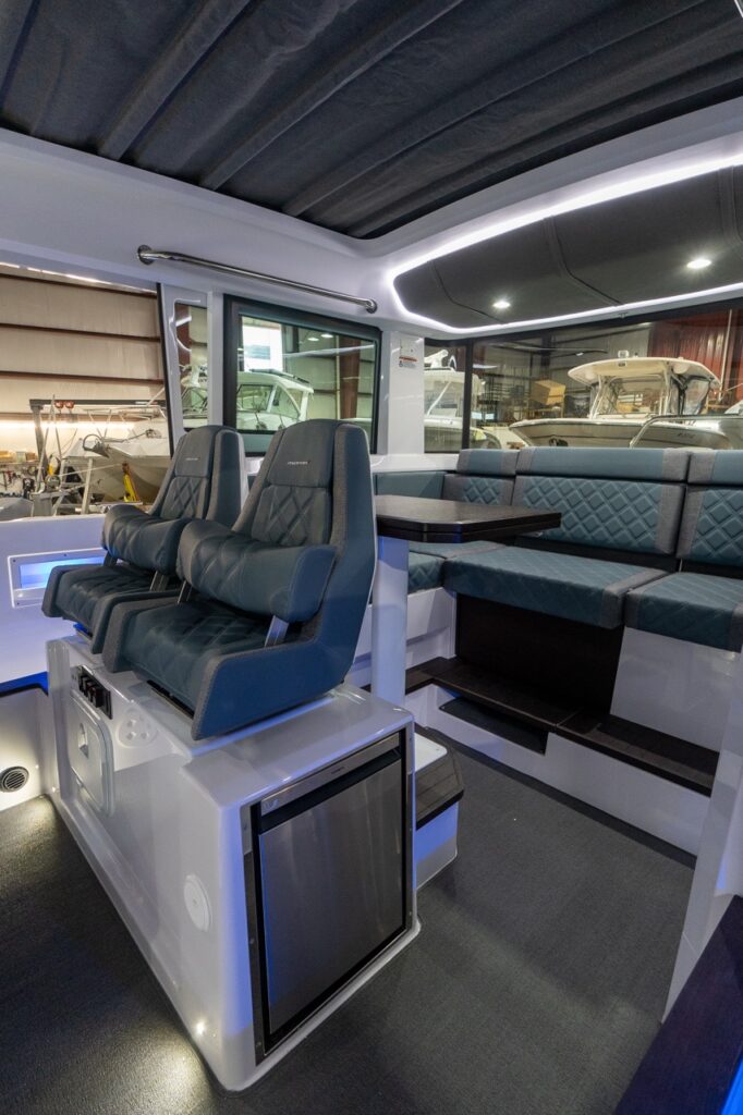 Inside the Axopar 37 XC pilothouse, two Petrol gray-blue helm seats sit above a stainless-front fridge module, with a dark-wood table and an aft bench seat with diamond-stitched Petrol upholstery under LED perimeter lighting.