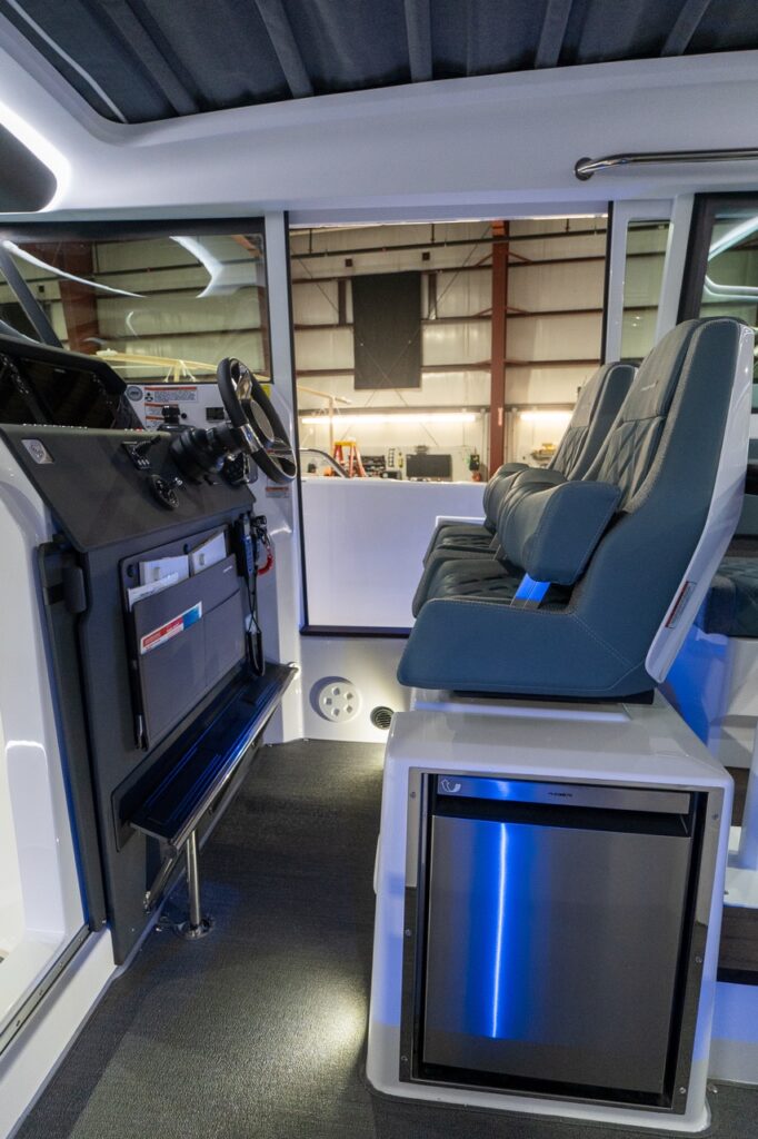 Side view inside the Axopar 37 XC pilothouse showing the driver console and steering wheel on the left, two Petrol gray-blue suspension seats on the right, and a stainless-front refrigerator built into the center module beneath the seats.