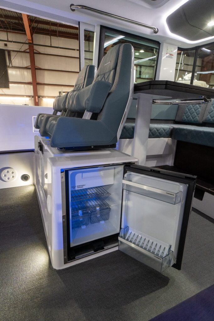 Inside the Axopar 37 XC pilothouse, the under-seat refrigerator is shown with its door open beneath two Petrol gray-blue suspension seats, revealing wire shelves and door bins next to the dinette table.