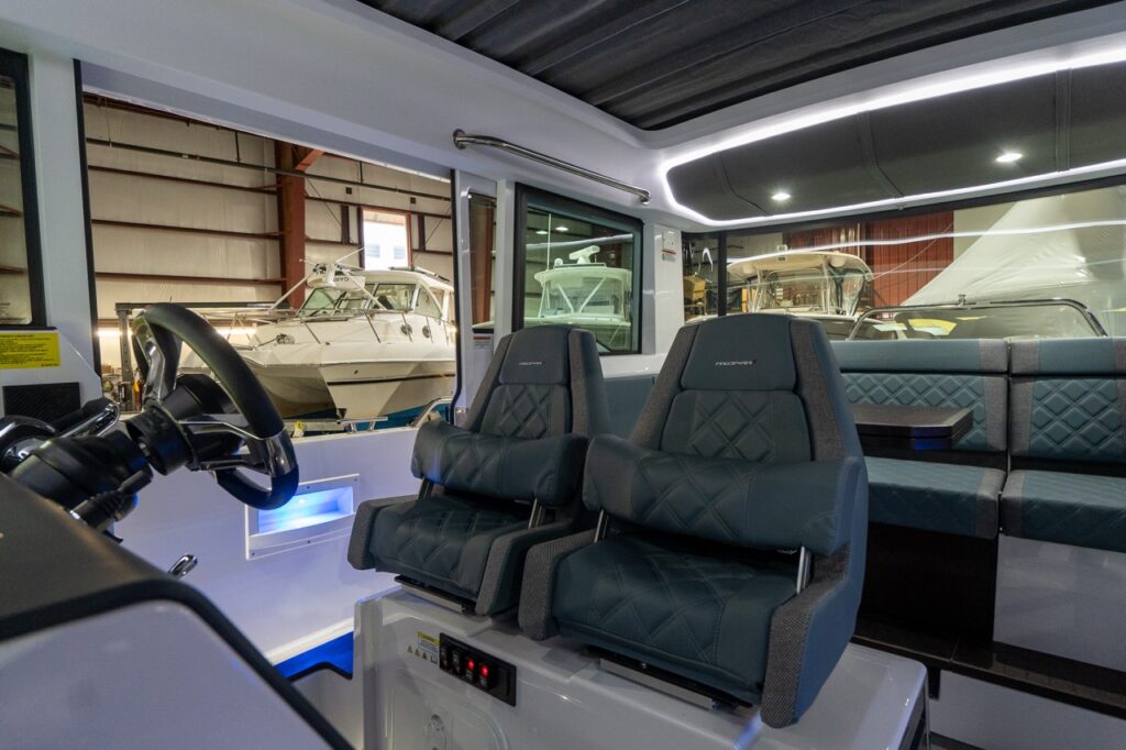 Inside the Axopar 37 XC pilothouse, two Petrol gray-blue suspension helm seats sit beside the steering wheel, with a matching Petrol upholstered aft dinette and table visible behind them under LED-lit ceiling panels.