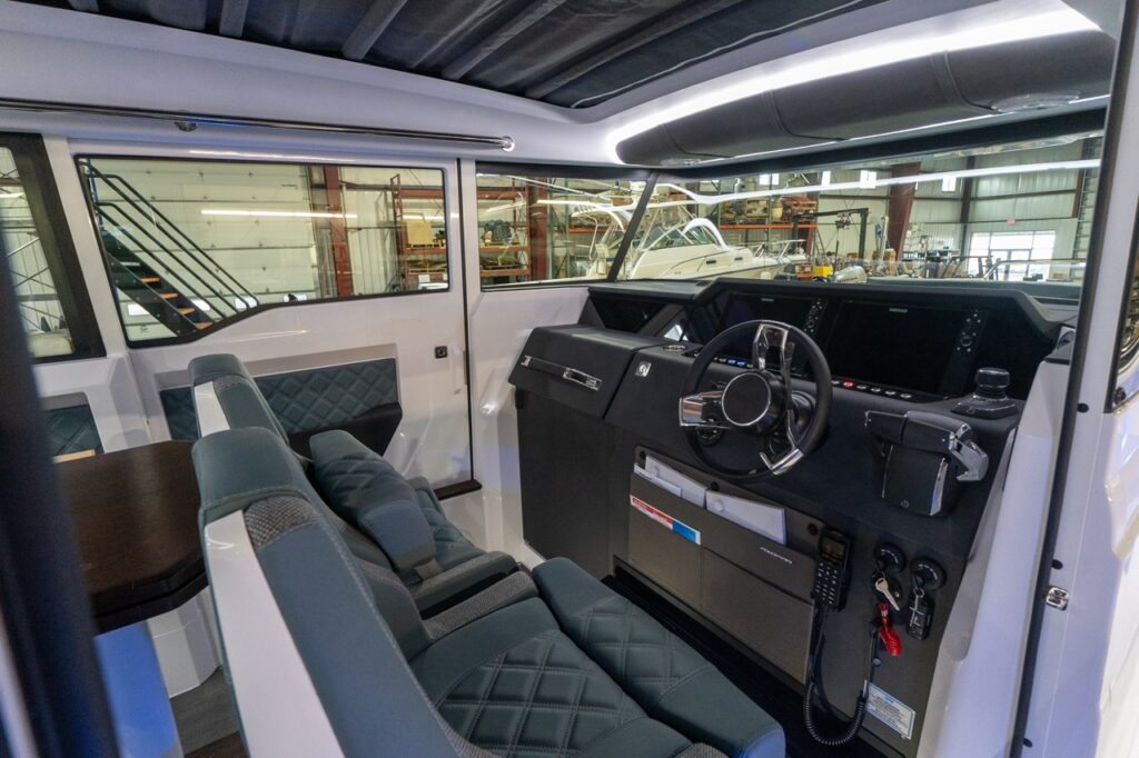 Angled view inside the Axopar 37 XC pilothouse showing the black helm console with steering wheel, dual Simrad displays, and controls beside two quilted Petrol gray-blue seats next to the dinette table.