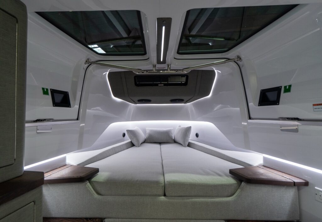 Forward-facing view of the Axopar 37 XC’s forward cabin showing a large V-berth with light upholstery and three pillows, twin overhead skylights, and LED accent lighting along the cabin sides with small wood-trim shelves at the foot of the berth.