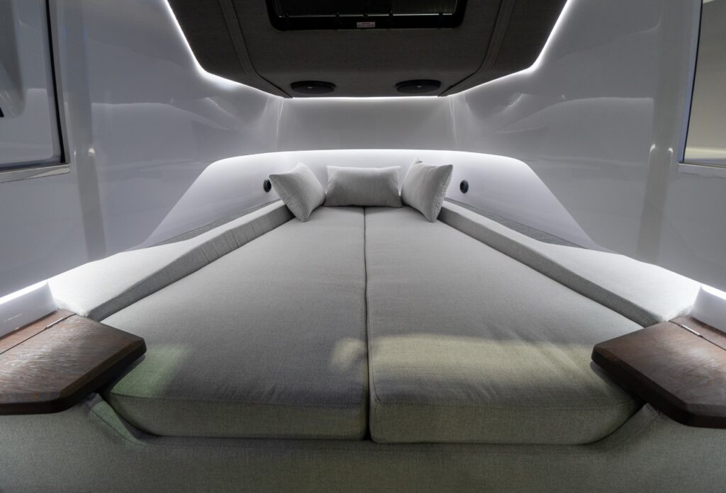 Close-up of the Axopar 37 XC forward cabin V-berth with two large light-gray cushions meeting at center, three pillows at the head, dark wood corner shelves, and soft LED strip lighting outlining the cabin walls and headliner.