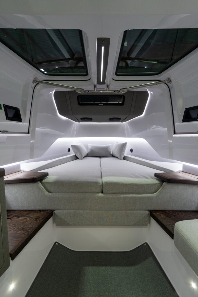 Symmetrical view of the Axopar 37 XC forward cabin showing a wide V-berth with light-gray cushions and three pillows, twin overhead skylights, a central LED ceiling light, and white fiberglass surfaces accented by continuous LED strip lighting and dark wood side shelves.