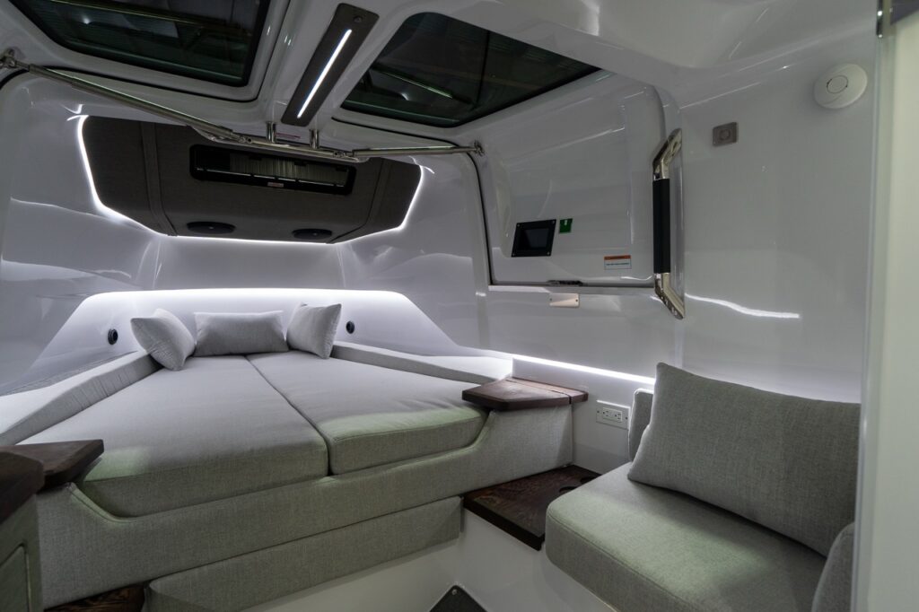 Angled view inside the Axopar 37 XC forward cabin showing the V-berth to the left with light-gray cushions and pillows, LED strip lighting along the hull sides, twin skylights overhead, dark wood corner shelves, and a small upholstered side seat with a back cushion on the right beside a charging outlet.