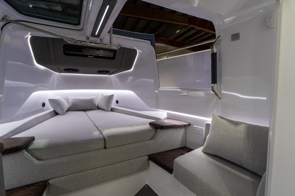 Inside the Axopar 37 XC forward cabin, the V-berth sits to the left with light-gray cushions and three pillows, while a small upholstered side seat is on the right. One overhead gullwing hatch is open, revealing the warehouse ceiling above, with LED strip lighting glowing along the cabin sides and dark wood corner shelves beside a charging outlet.