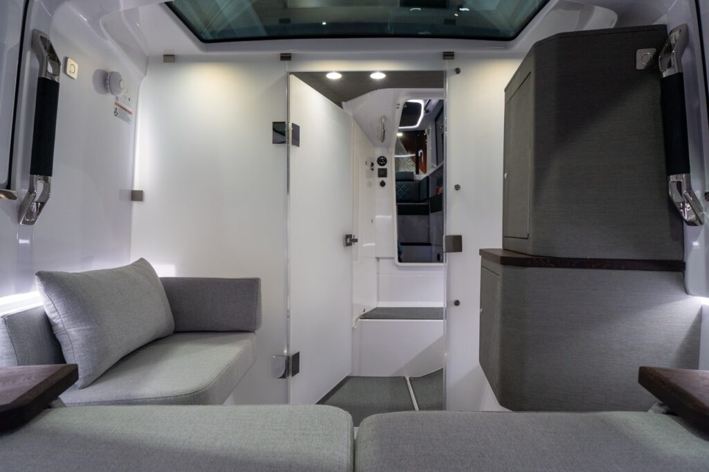 View from the forward cabin looking aft toward the pilothouse: a white privacy door is swung open across the passage to enclose the head compartment, while the walkway steps lead up toward the cabin entry. To the left is a compact gray upholstered lounge seat with a cushion, and to the right are two tall fabric-faced storage cabinets with a dark wood trim shelf. Overhead skylights and recessed lights brighten the white interior.