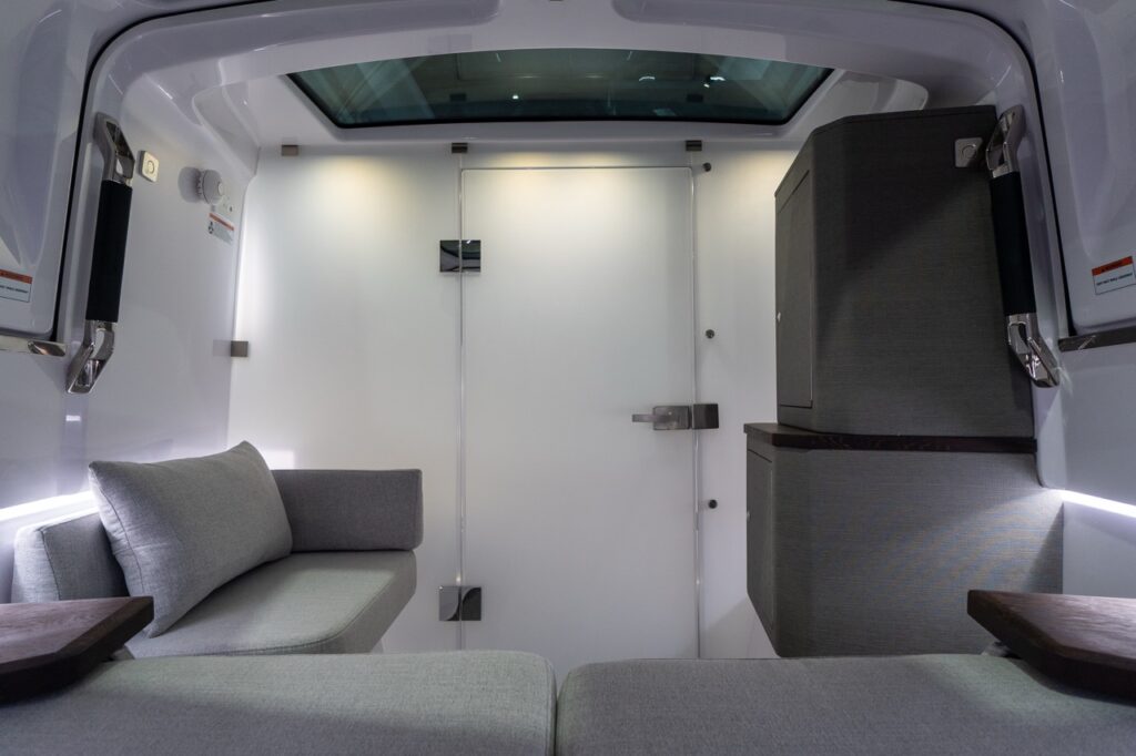 Inside the forward cabin, a white privacy door is fully closed beneath a large overhead skylight. A compact gray upholstered lounge with a matching cushion sits to port, while two tall fabric-faced storage cabinets with a dark wood trim ledge are to starboard. LED accent lighting runs along the cabin sides, highlighting the clean, bright interior.